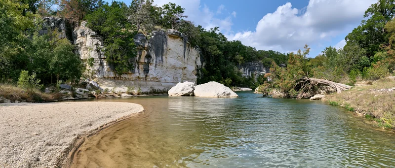 Shallow clear river flowing alongside sandy gravel bank with limestone cliffs and trees under blue sky