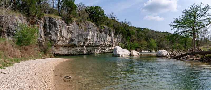 Clear shallow river with sandy gravel bank curving alongside limestone cliff under blue sky