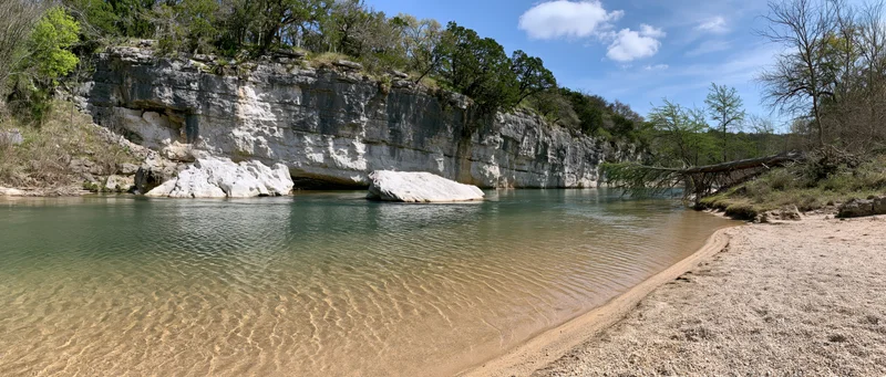 Clear shallow river with sandy gravel bank curving alongside rocky cliff under blue sky