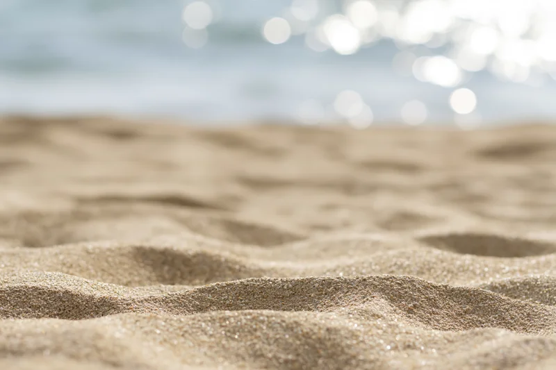 Close-up of textured sand on a beach with blurred ocean and sunlight in the background