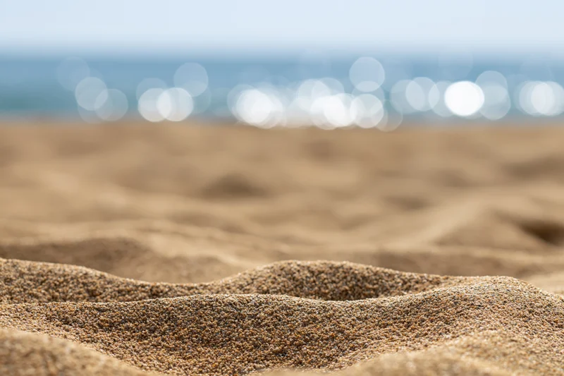 Close-up of textured golden sand on a beach with blurred ocean and bokeh lights in the background