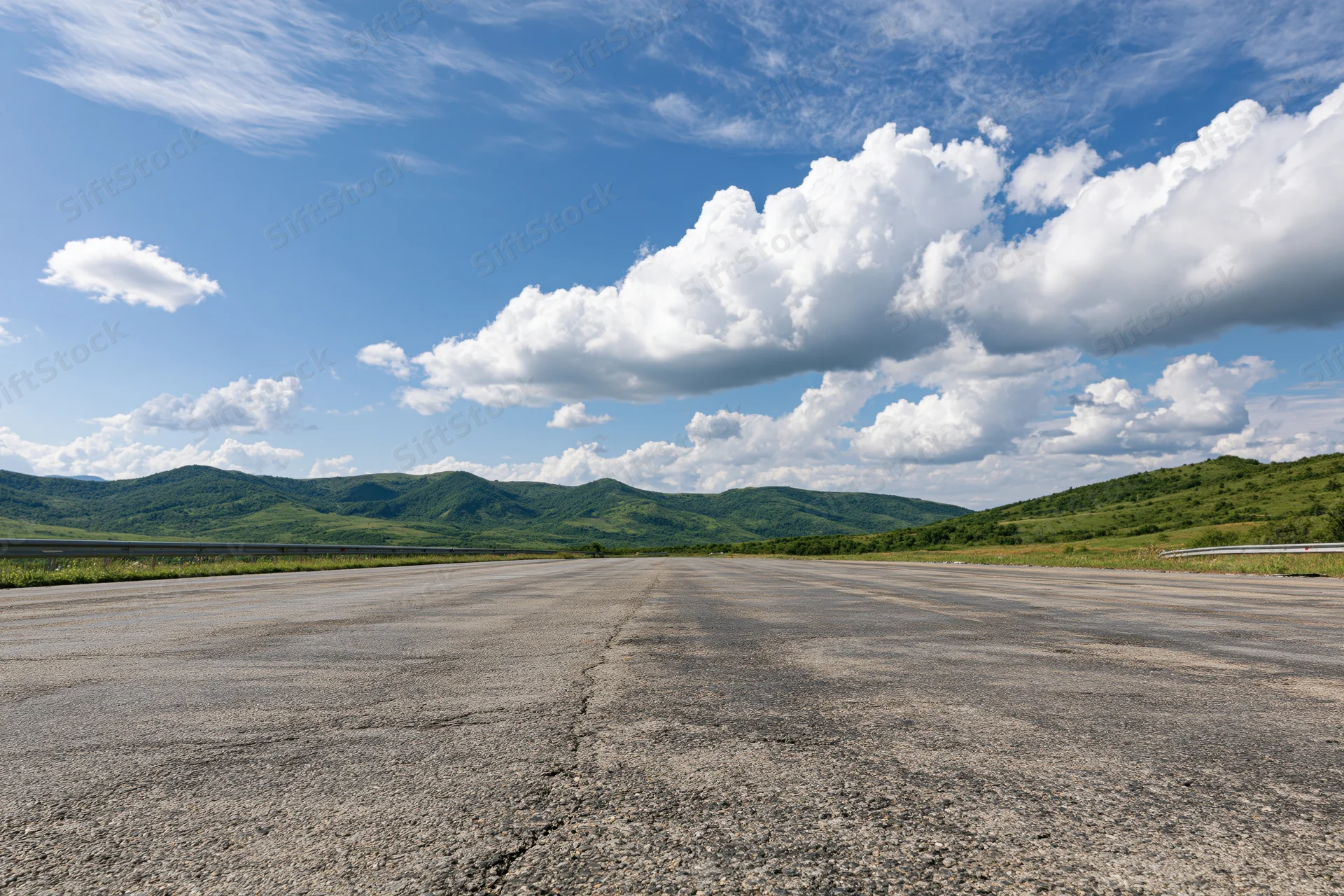 Empty asphalt highway stretching to the horizon under a blue sky with white clouds, flanked by green hills and mountains