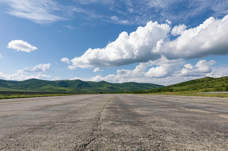 Empty asphalt highway stretching to the horizon under a blue sky with white clouds, flanked by green hills and mountains