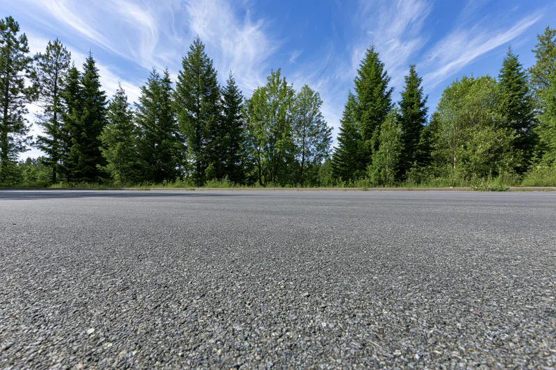 Empty asphalt road surface stretching toward a tree line under a blue sky with wispy clouds
