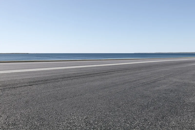 Empty asphalt road with white lane markings running parallel to the ocean under a clear blue sky