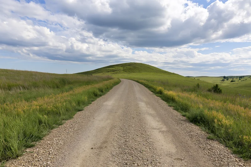 Gravel road stretching straight toward a rounded green hill in a grassy rural landscape