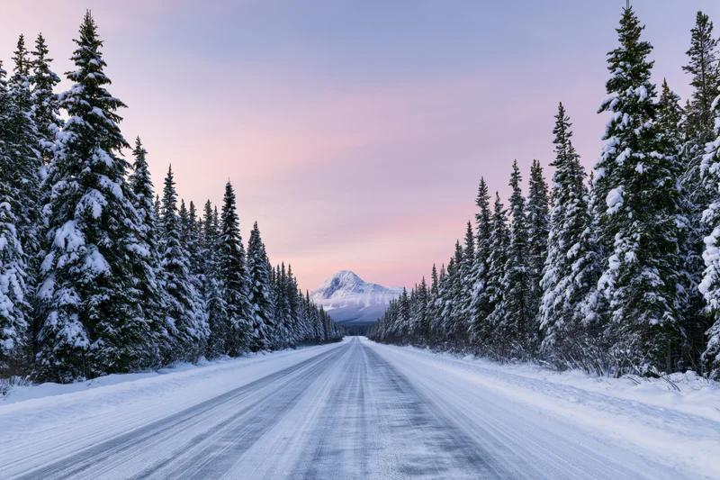 Snow-covered highway stretching through evergreen forest toward distant mountain at twilight