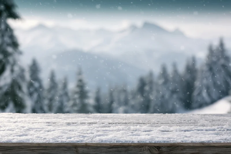 Snow-dusted wooden table in front of blurred winter mountain landscape with pine trees
