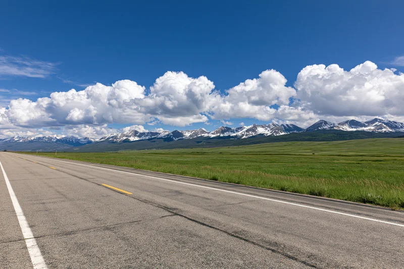 Empty two-lane asphalt highway stretching across a green field toward snow-capped mountains under a blue sky with white clouds