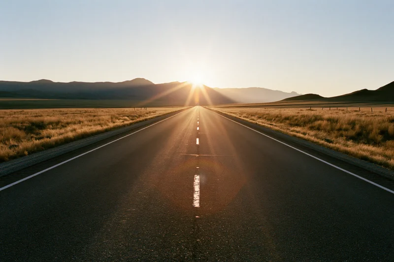 Straight asphalt highway stretching toward the sun at sunset with mountains on the horizon