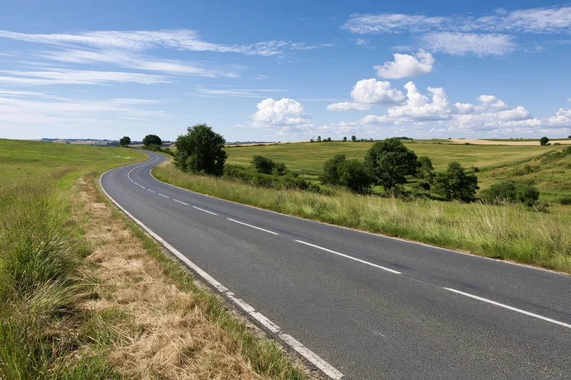Winding two-lane asphalt road through green rolling countryside under blue sky with clouds