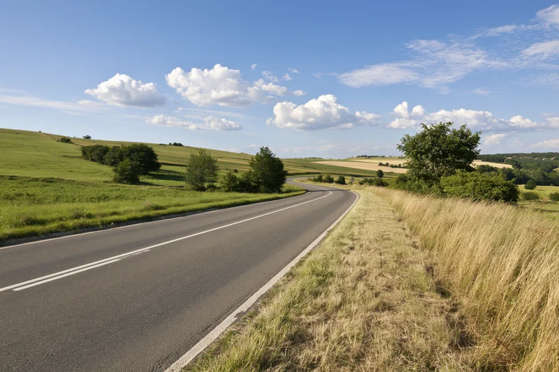Winding two-lane asphalt road through green rolling countryside under blue sky with white clouds
