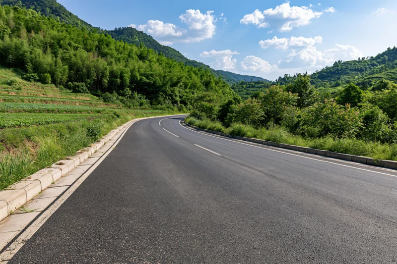 Winding asphalt road curving through green hills and farmland under a blue sky with white clouds