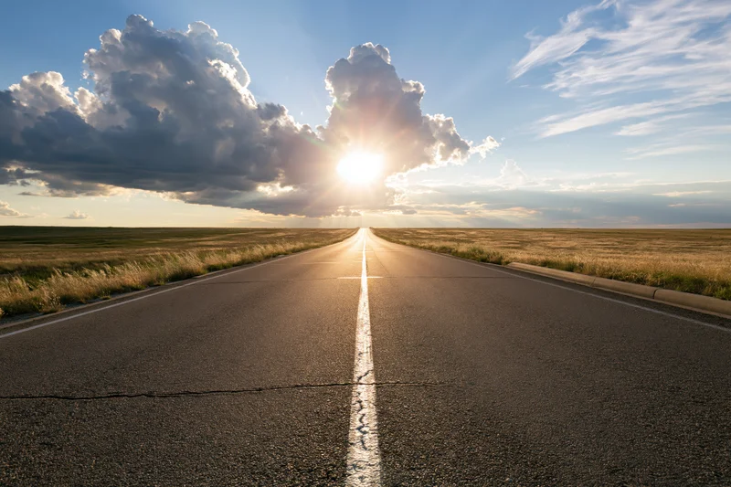 Straight asphalt road stretching to the horizon under a sunlit sky with clouds and golden light