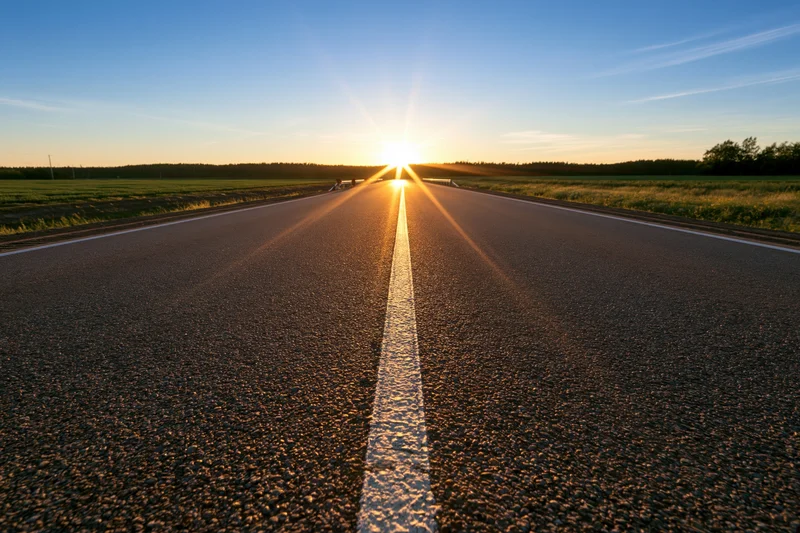 Straight asphalt road stretching toward the horizon at sunset with white center line and fields on either side