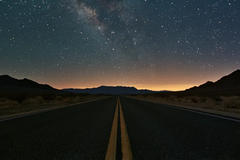Straight two-lane highway stretching into the desert night under a star-filled sky with mountains on the horizon