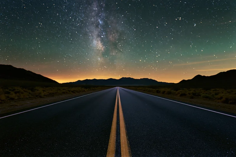 Straight desert highway under starry night sky with milky way above distant mountains