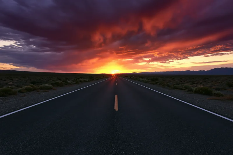 Straight two-lane highway stretching to the horizon under a fiery sunset sky in the desert