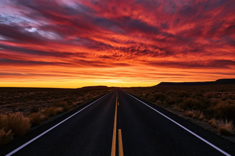Straight two-lane highway stretching into the desert horizon under a fiery sunset sky