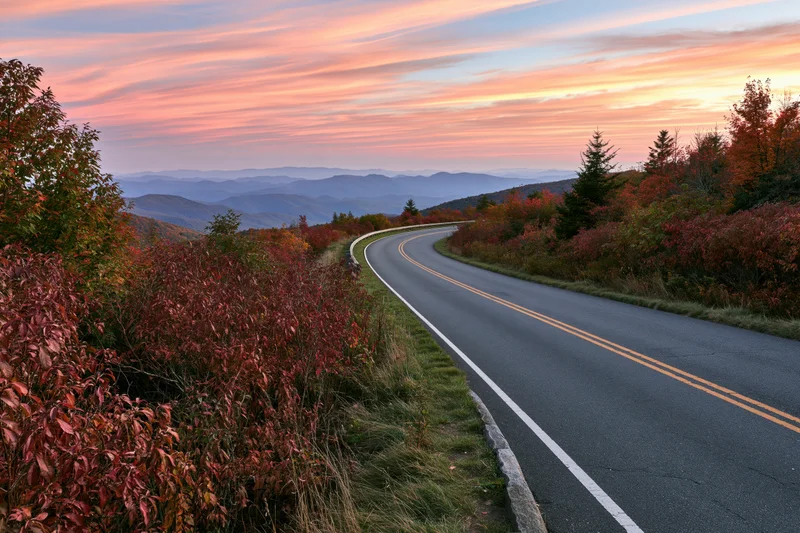 Winding two-lane highway through autumn forest at sunset with mountain views