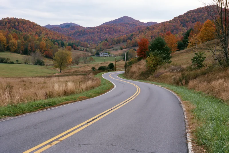 Winding two-lane rural highway through autumn countryside with colorful foliage and distant mountains