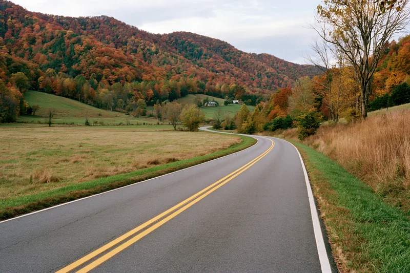 Winding two-lane asphalt road through autumn countryside with colorful forested hills and open fields