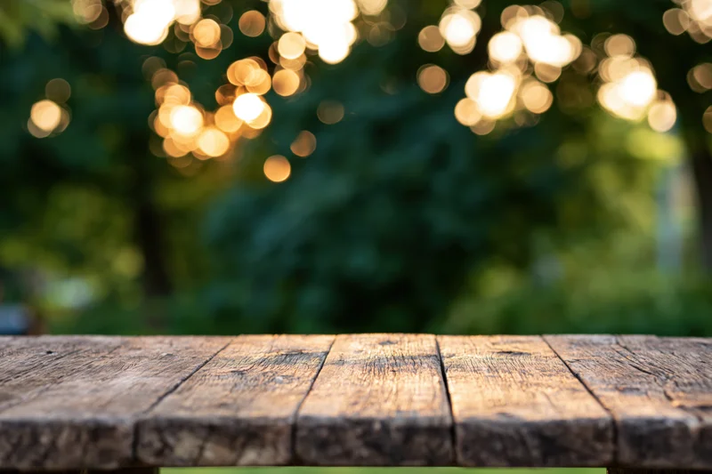 Weathered wooden table surface in a garden with blurred green foliage and warm bokeh lights in the background