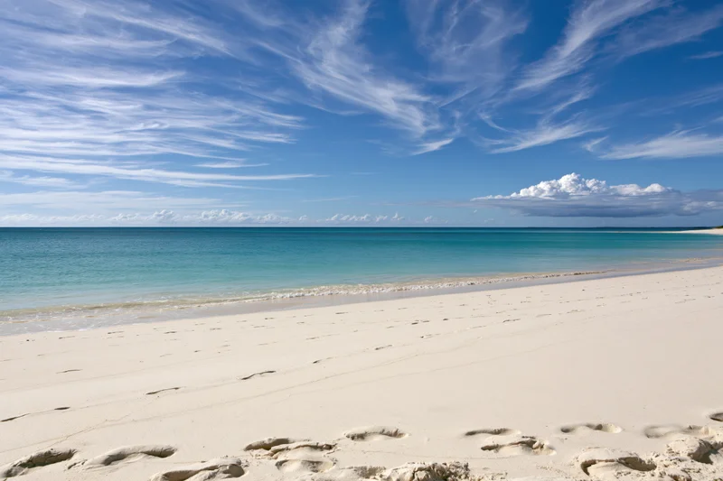 White sand beach with footprints leading to calm turquoise ocean under blue sky with wispy clouds