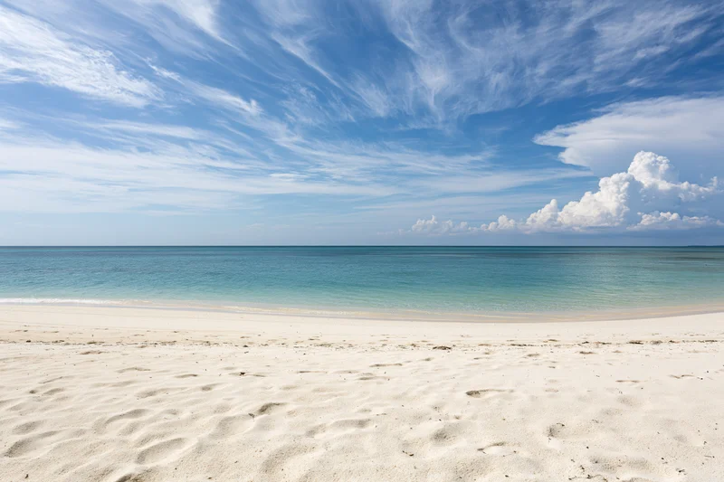 White sand beach stretching to calm turquoise ocean under blue sky with wispy clouds