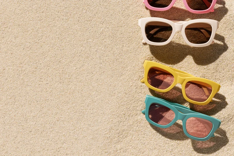 Four colorful retro-style sunglasses arranged vertically on sandy beach surface