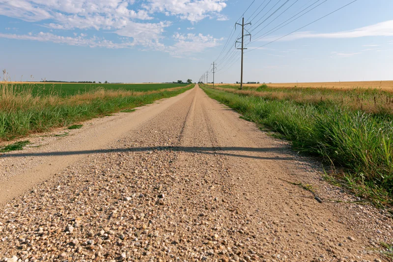 Gravel road stretching straight to the horizon between green and golden fields under a blue sky with scattered clouds
