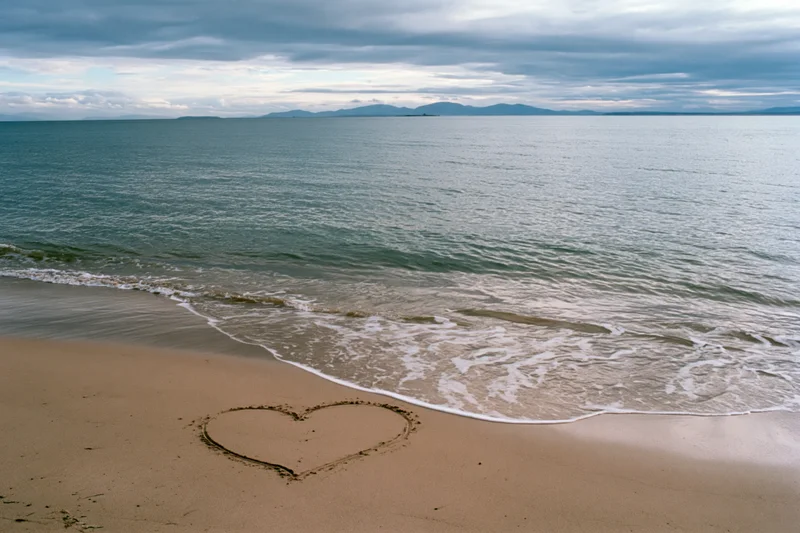 Heart shape drawn in wet sand on a quiet beach with gentle waves and distant mountains under cloudy sky