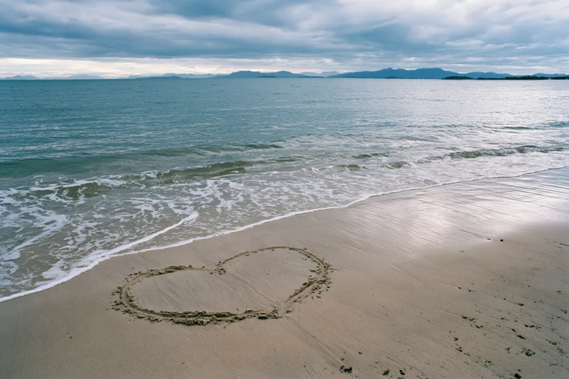 Heart drawn in wet sand on a quiet beach with gentle waves and distant mountains under overcast sky