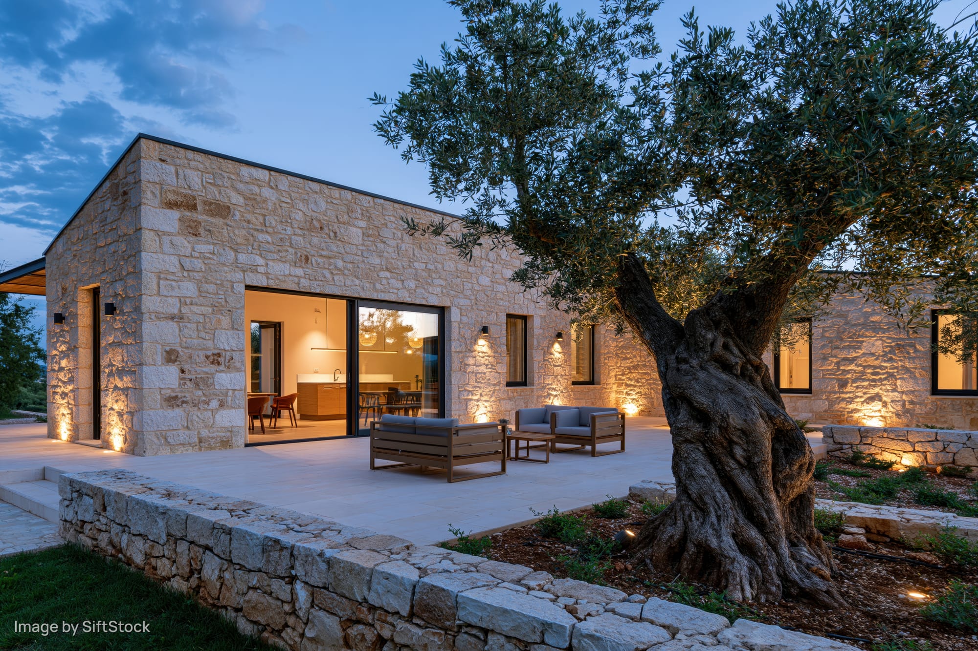 Stone villa exterior at dusk with illuminated courtyard and olive tree