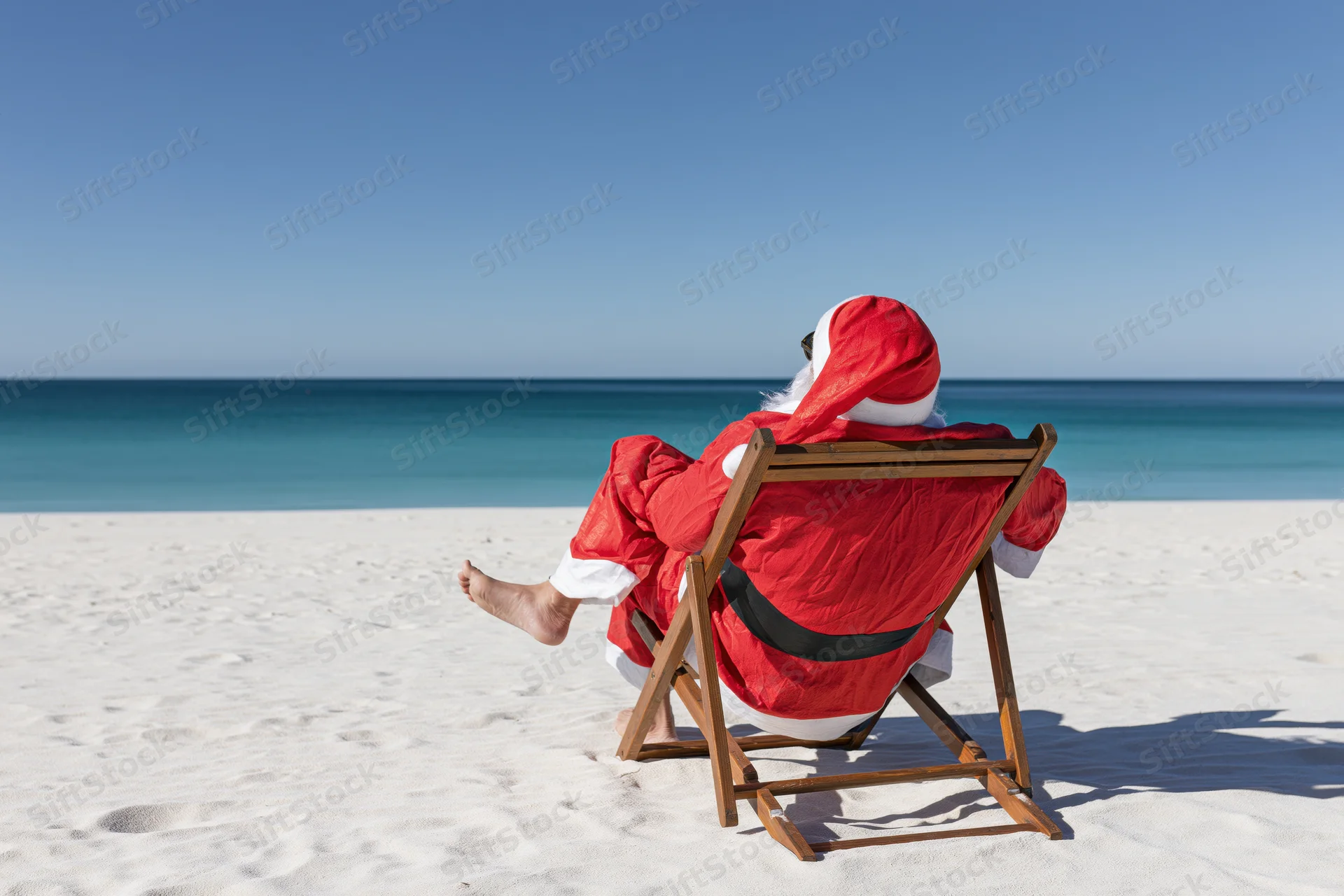 Santa reclining in a wooden deckchair on a sandy beach facing the ocean