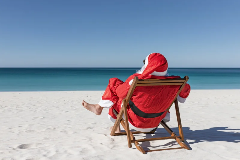 Santa reclining in a wooden deckchair on a sandy beach facing the ocean