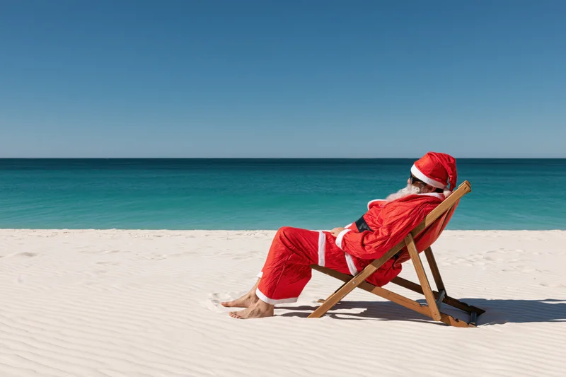 Santa reclining in a deckchair on a sandy beach beside the ocean