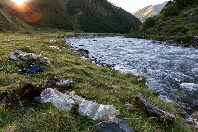 Plastic bottles and bags scattered along a grassy riverbank beside a flowing river in a mountain valley at sunset