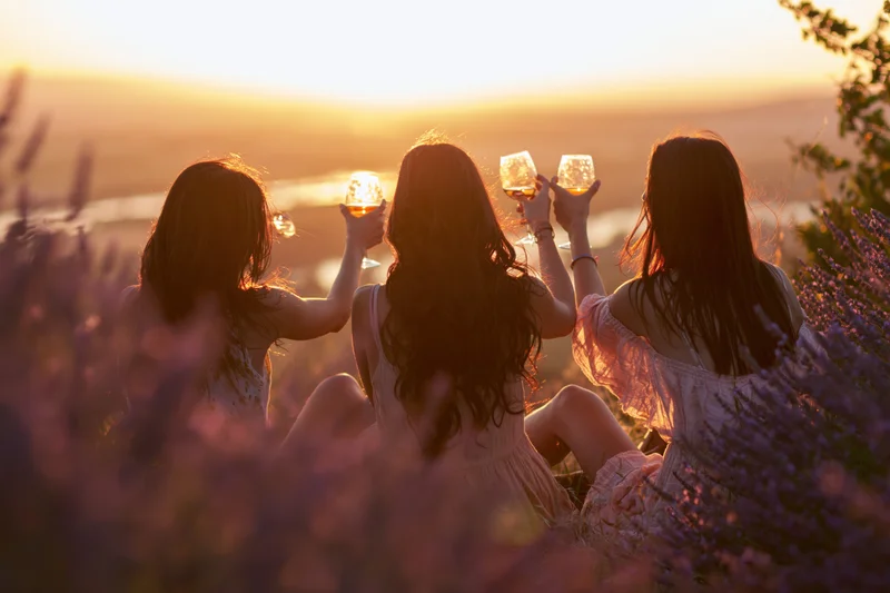 Three women toasting with wine glasses in a lavender field at sunset