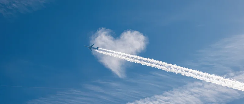 Airplane flying away from heart-shaped cloud in blue sky leaving contrail