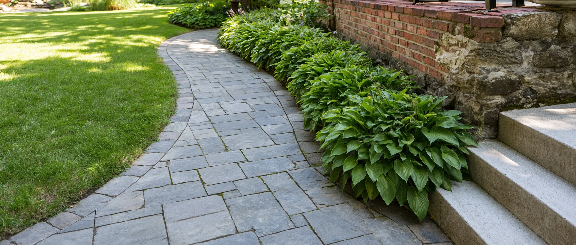 Curved stone pathway winding through a garden bordered by hosta plants and brick wall