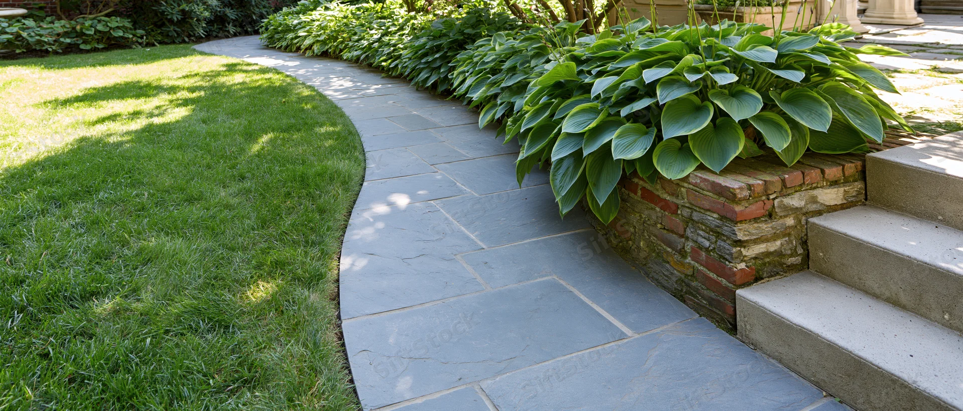 Curved stone pathway winding through a green lawn bordered by hosta plants and brick retaining wall near steps