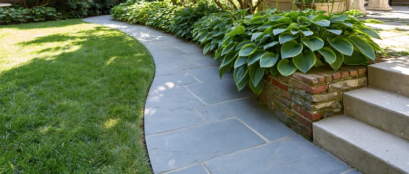 Curved stone pathway winding through a green lawn bordered by hosta plants and brick retaining wall near steps