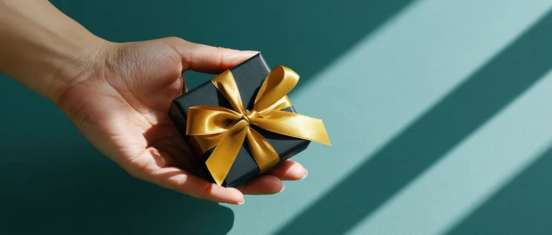 Hand holding small black gift box with gold ribbon against teal background