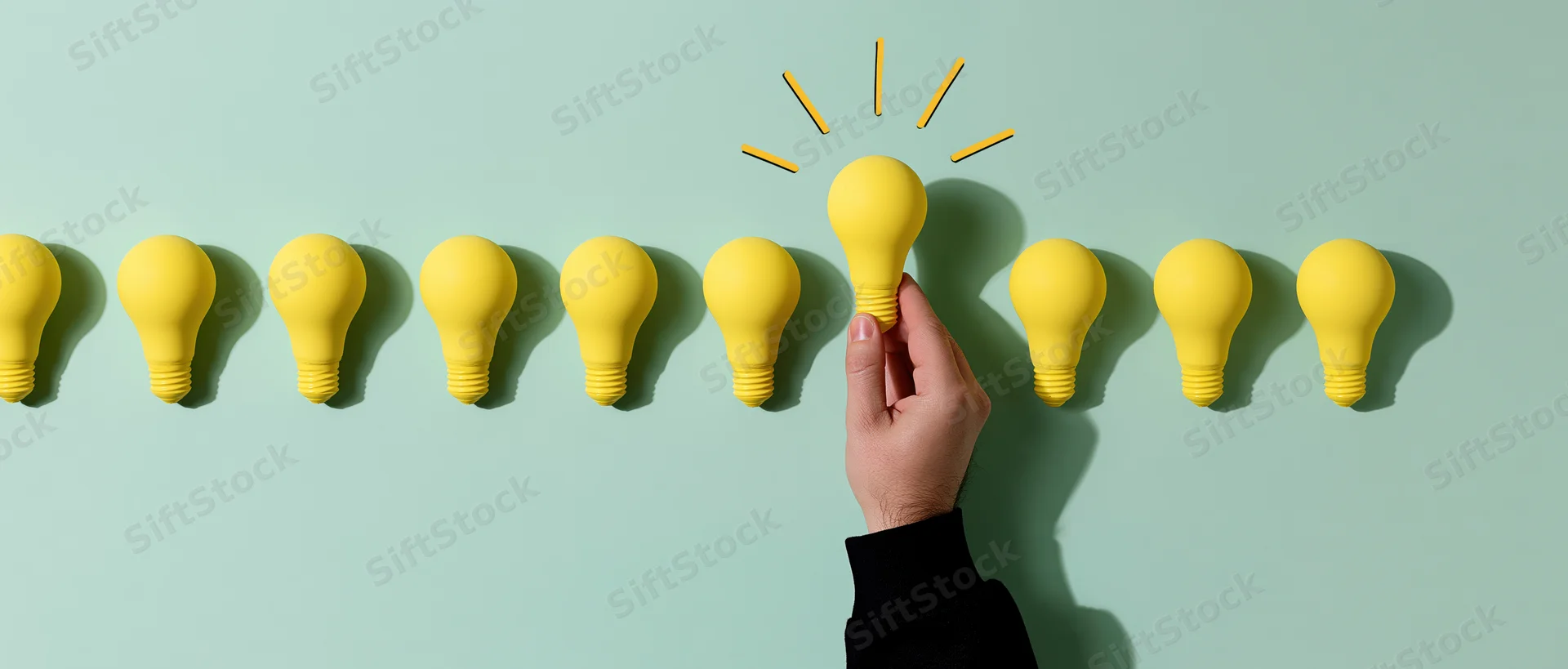 Hand lifting one yellow lightbulb from a row against a mint green background