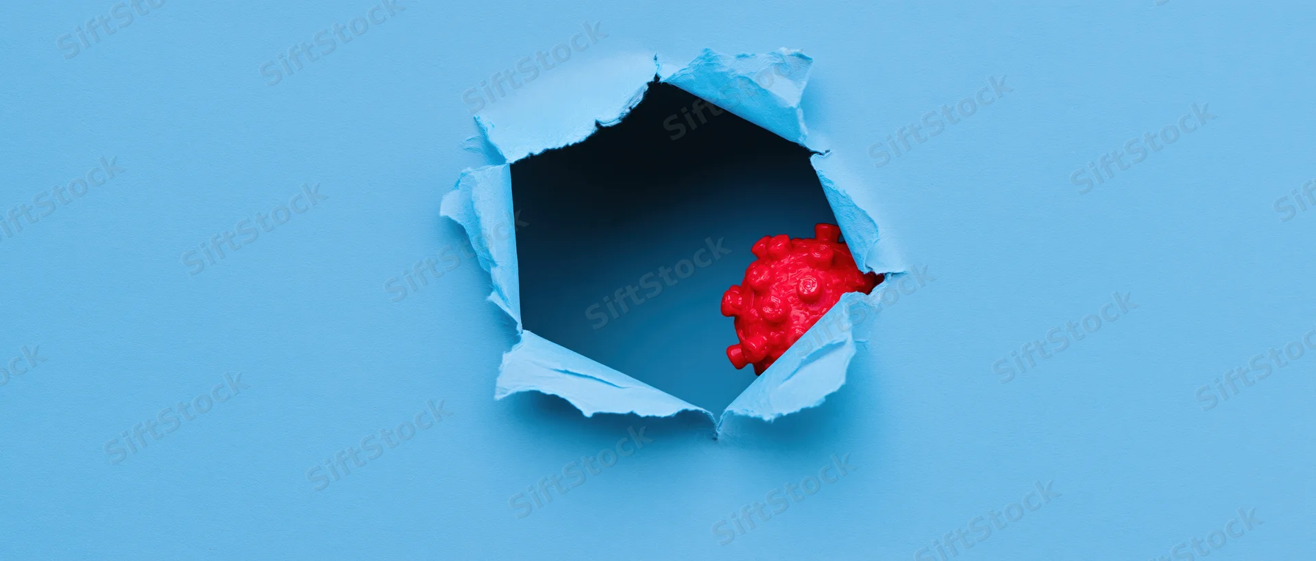 Red plastic coronavirus model emerging from a torn hole in blue paper
