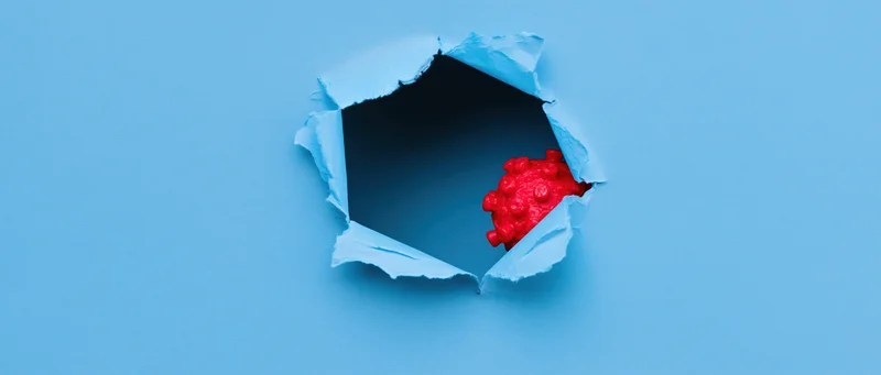 Red plastic coronavirus model emerging from a torn hole in blue paper