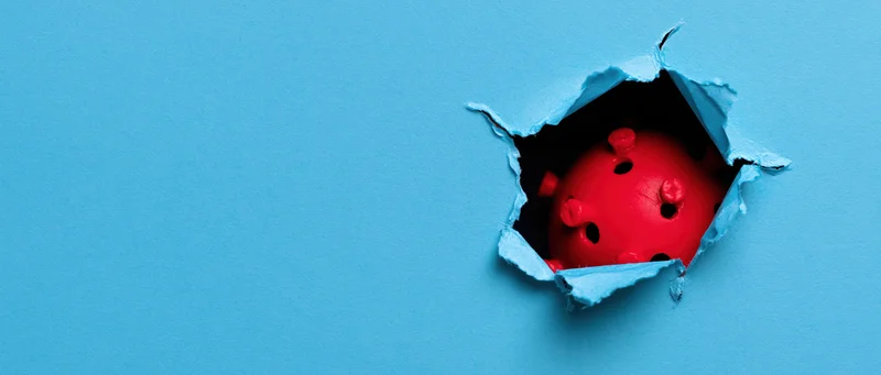 Red plastic coronavirus model emerging through torn blue paper
