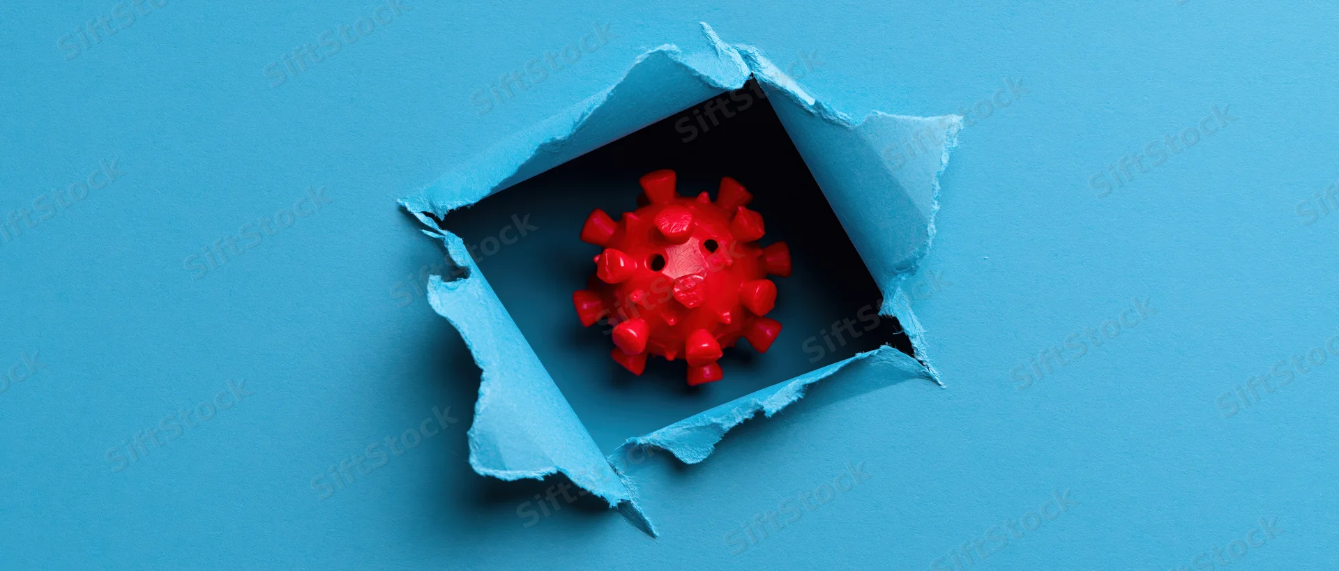 Red plastic coronavirus model visible through a torn hole in blue paper