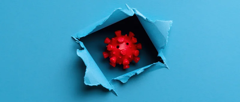 Red plastic coronavirus model visible through a torn hole in blue paper